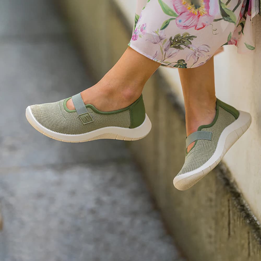 Olive knit shoes with a strap, worn with a floral dress on a stone ledge.