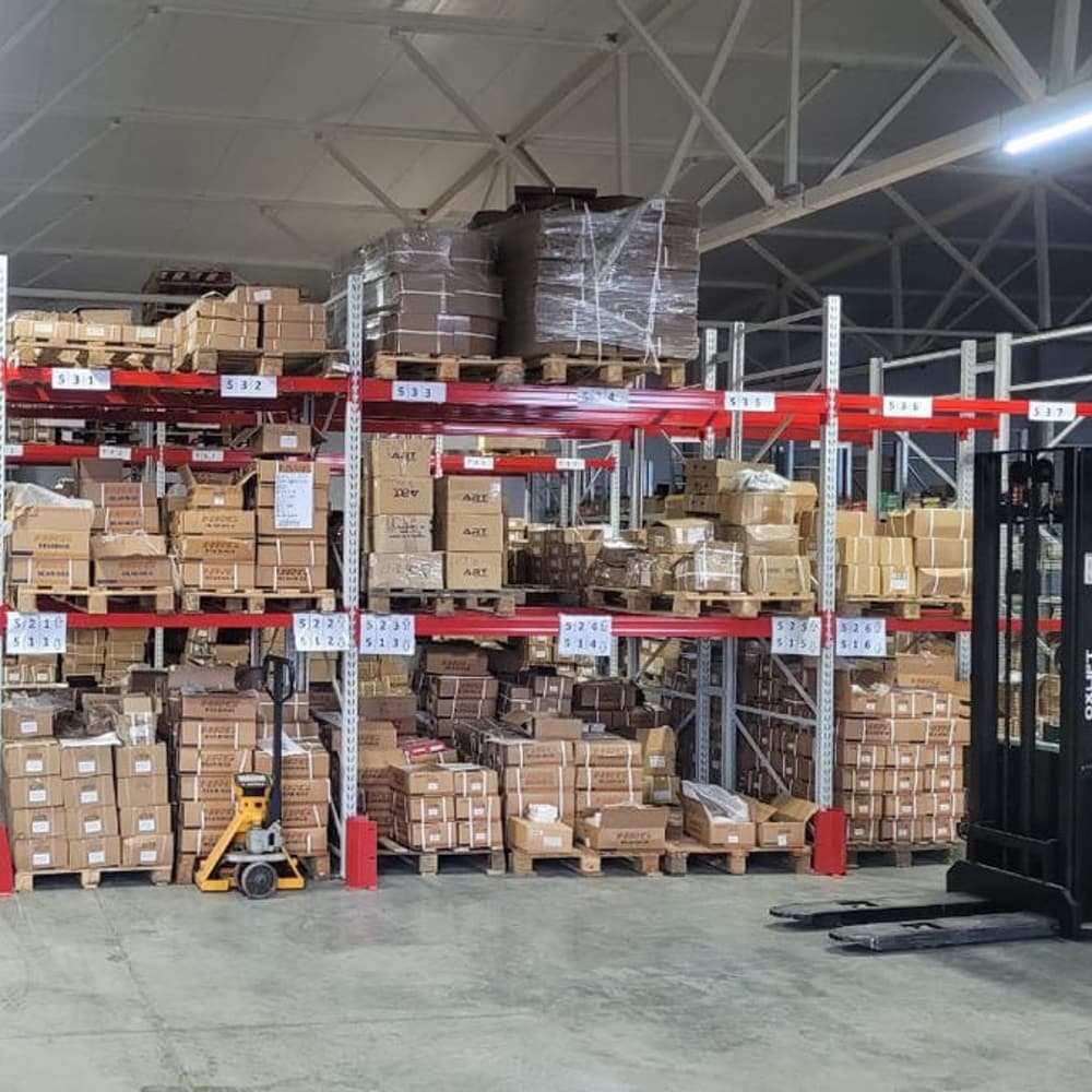 A warehouse hall: racking loaded with pallets of cartons, a pallet truck and a forklift on the floor.